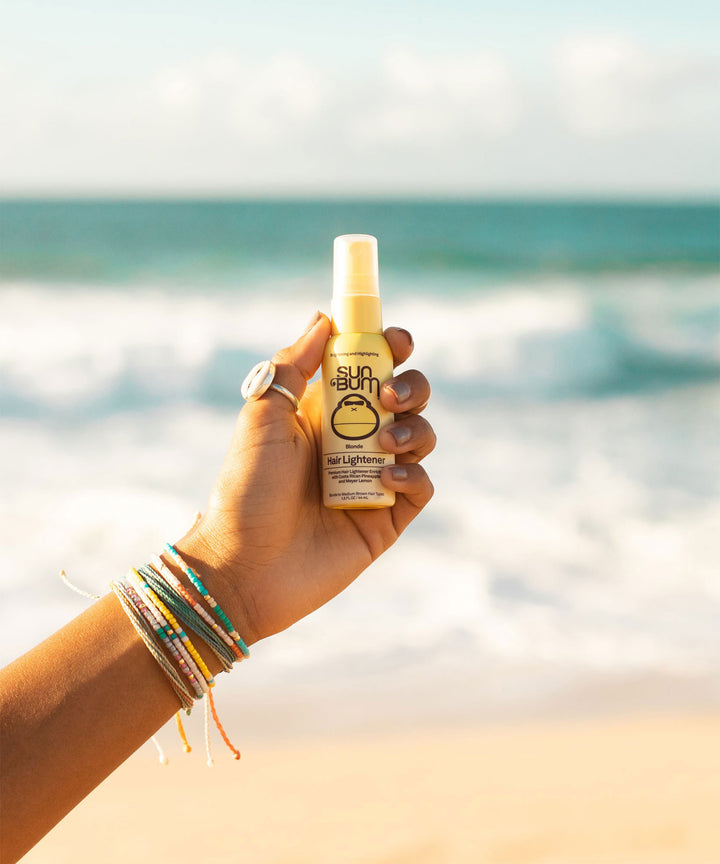 Hand holding Sun Bum Blonde Hair Lightener spray bottle against a beachy ocean backdrop, adorned with colorful friendship bracelets, showcasing the natural hair lightening product by Sun Bum