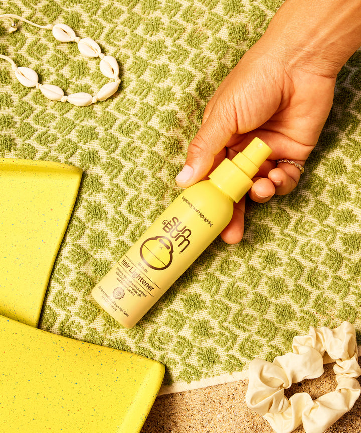 A hand holding a yellow Blonde Hair Lightener spray bottle against a green patterned background, with yellow accessories and shell necklace decoratively arranged, by Sun Bum.