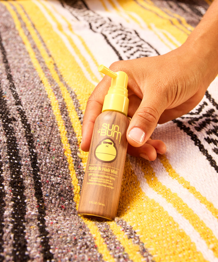 A hand holding Sun Bum's Scalp & Hair Mist SPF 30 spray bottle against a yellow and black patterned beach towel background, showcasing the lightweight sun protection product for hair - by Sun Bum