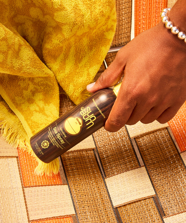 A hand holding Sea Spray texturizing spray bottle against warm-toned woven mat backdrop with yellow beach towel, showcasing Sun Bum's beach-inspired hair styling product in natural lighting - by Sun Bum