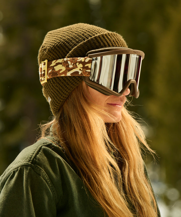 Stylish ski goggles featuring mirrored lens with camouflage strap design, paired with olive knit beanie, shown in warm outdoor lighting - Sun Bum X Electric Goggles collection for premium mountain eye protection from Sun Bum