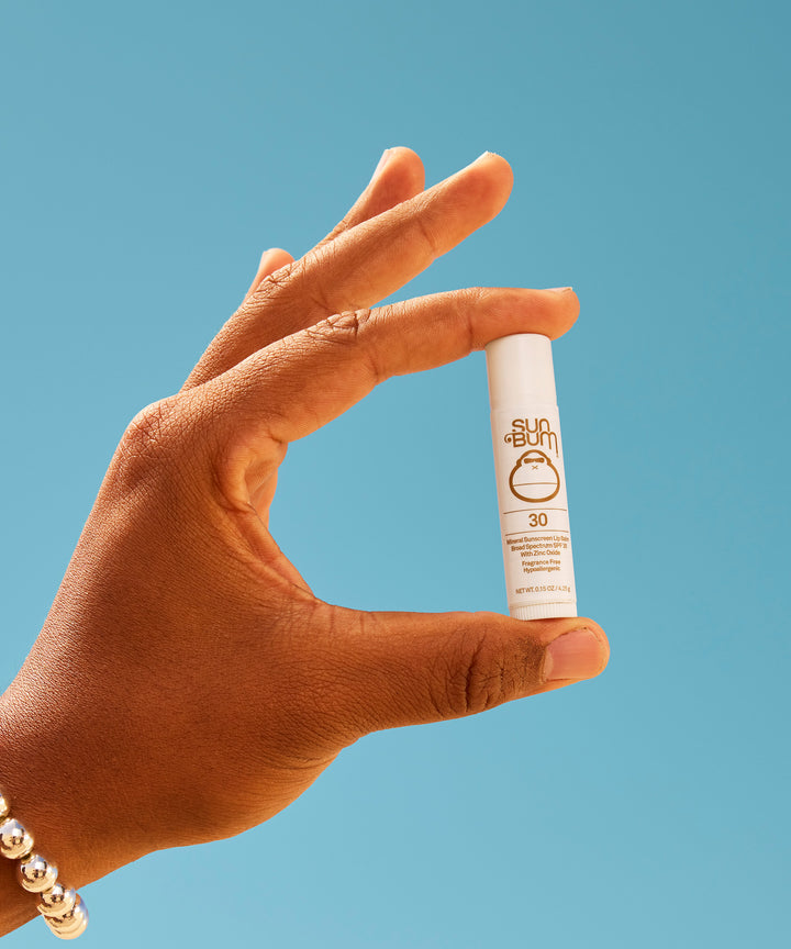 Hand holding Mineral SPF 30 Sunscreen Lip Balm against blue background, showing white cylindrical tube with zinc-based protection formula - by Sun Bum