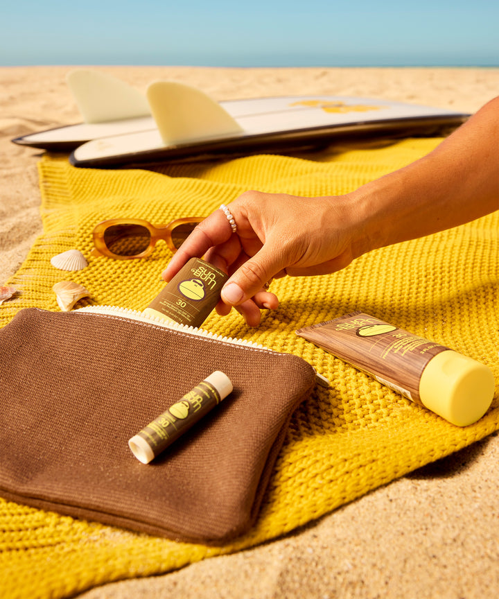 Beach essentials on yellow towel with surfboard, sunglasses and Road Tripper travel products from Sun Bum