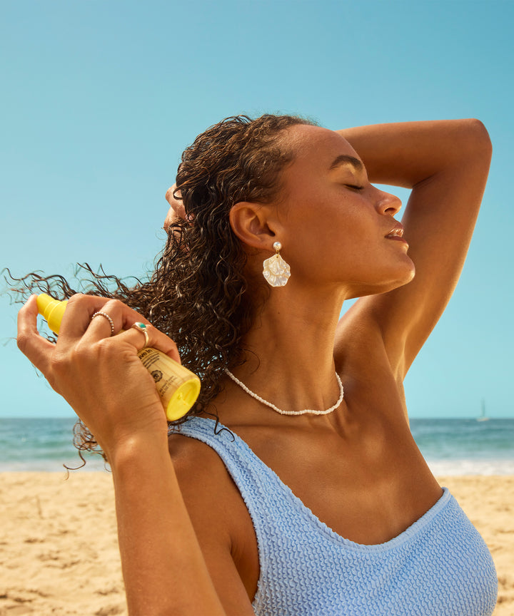 Model with curly hair applying 3 In 1 Leave In hair treatment at the beach, wearing a blue swimsuit and pearl jewelry against a sunny ocean backdrop with natural lighting showcasing the product application - Sun Bum