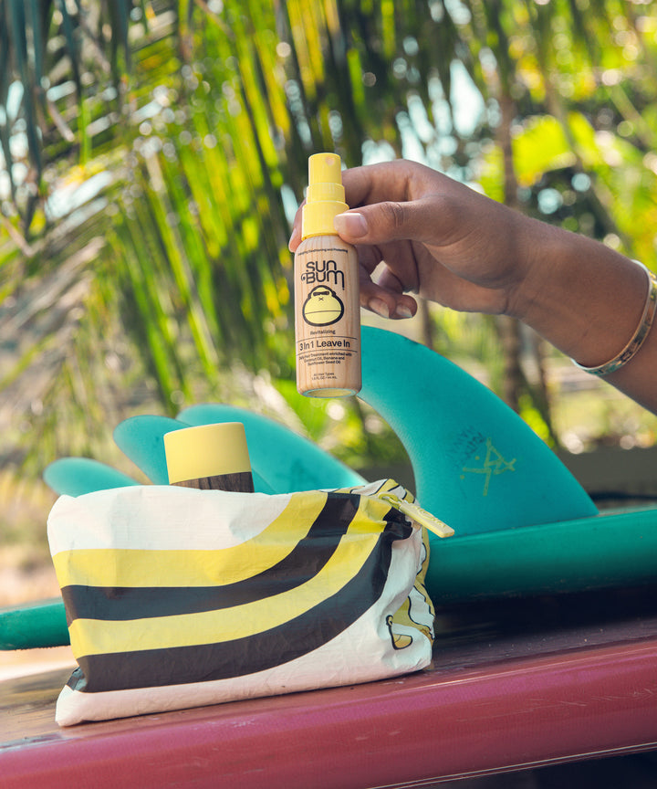A hand holding a bottle from the Hair Care Travel Essentials Kit next to a custom SPLASH-PROOF pouch by ALOHA Collection, with surfboards and tropical greenery in the background. Sun Bum.