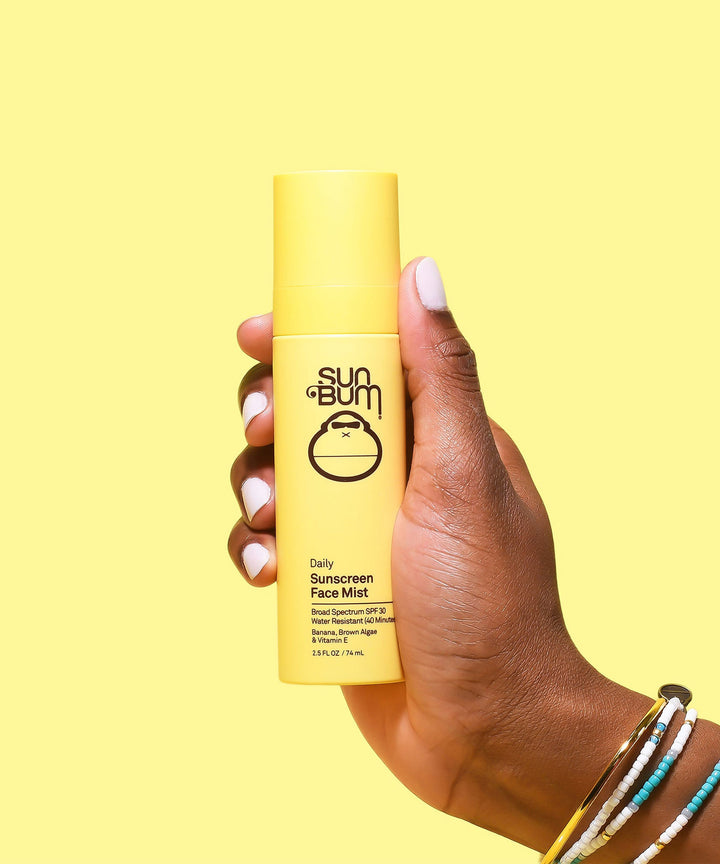 Hand holding a yellow bottle of Daily Sunscreen Face Mist SPF 30 against a bright yellow background. Product is formulated with antioxidant-rich Banana, Brown Algae, and Vitamin E. Sun Bum.