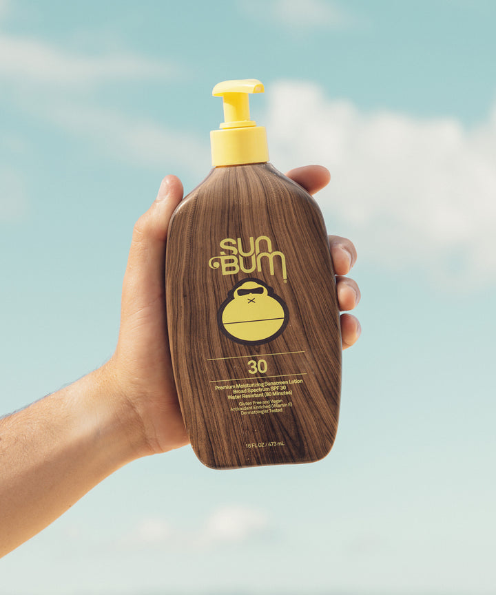 Hand holding a bottle of Original SPF 30 Sunscreen Lotion with a wooden texture and yellow pump against a clear sky, Sun Bum.