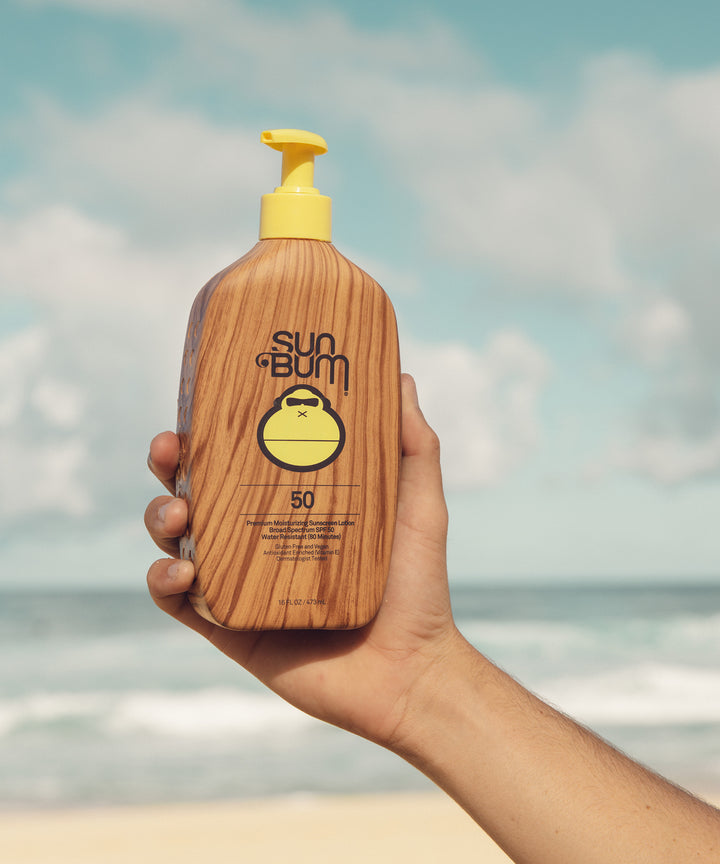 Hand holding a bottle of Original SPF 50 Sunscreen Lotion by the beach, Sun Bum