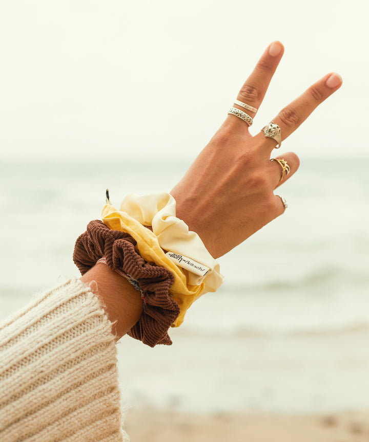 Person at the beach wearing a Sun Bum x Kitsch Limited Edition Scrunchie Set in yellow, brown, and cream, making a peace sign with their hand.