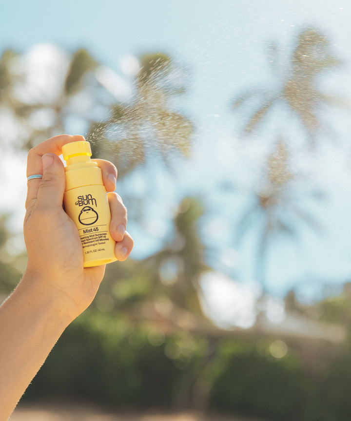 Hand holding a bottle of Original SPF 45 Sunscreen Face Mist being sprayed outdoors with palm trees in the background, Sun Bum.