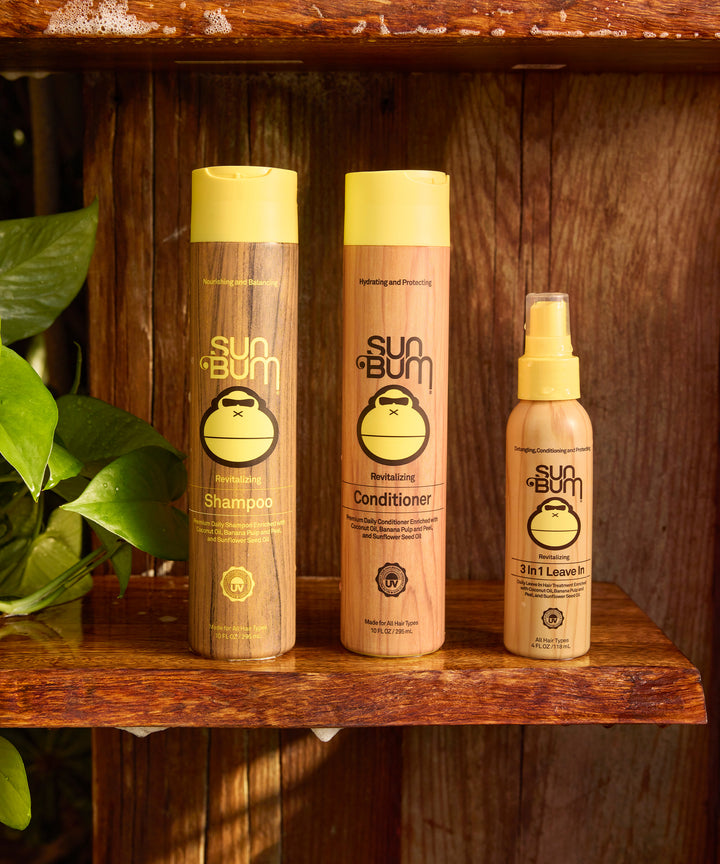 Three Sun Bum hair care products on a rustic wooden shelf - shampoo, conditioner, and 3 In 1 Leave In spray bottles with yellow caps and monkey logo, displayed alongside green plant leaves. - Sun Bum