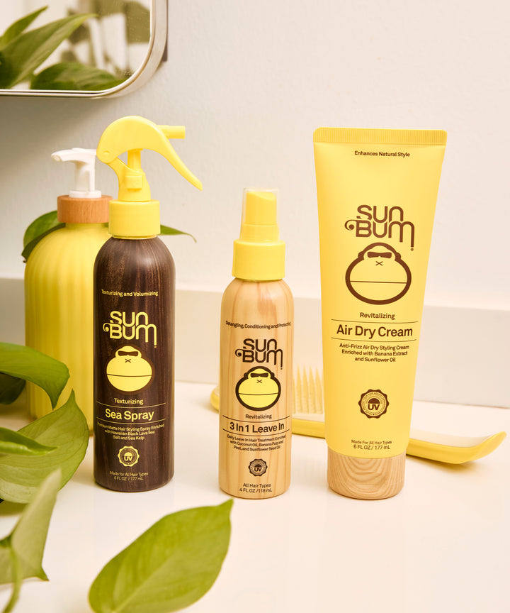 Yellow tube of Revitalizing Air Dry Cream displayed alongside other Sun Bum hair care products, including Sea Spray and 3-in-1 Leave In, set against a clean white background with green plant leaves for decoration - by Sun Bum