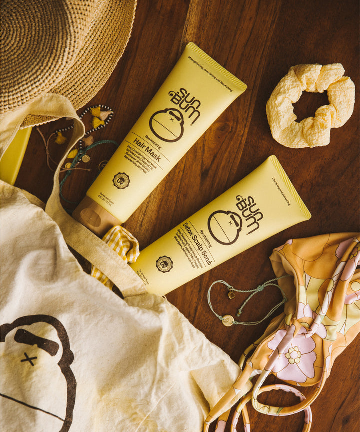 Yellow tubes of Revitalizing Detox Scalp Scrub displayed on wooden surface with straw hat, canvas bag, and accessories, showcasing the natural hair care product by Sun Bum