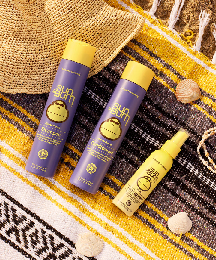 Blonde Hair Lightener spray bottle alongside matching purple shampoo and conditioner bottles displayed on a yellow striped beach blanket with seashells, showcasing Sun Bum's beach-inspired hair care collection. Perfect summer beach day styling products by Sun Bum.