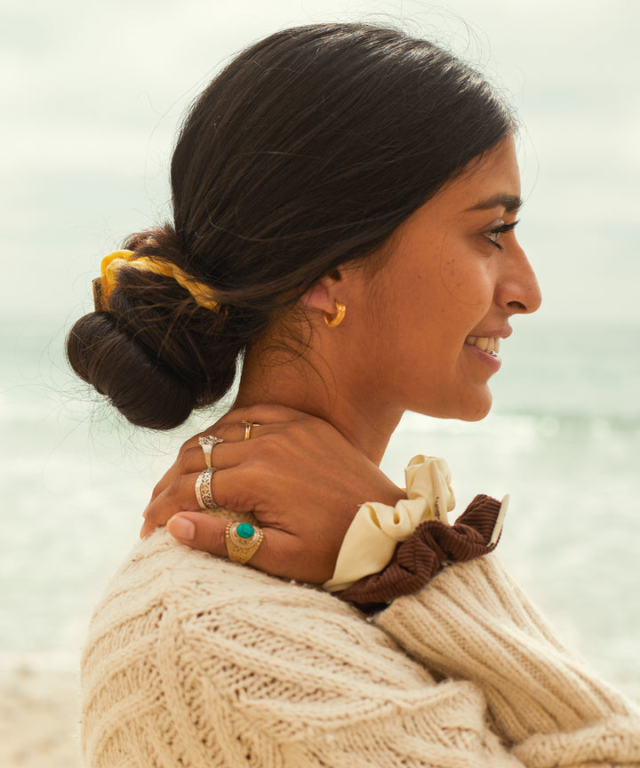 Woman wearing SB x Kitsch Limited Edition Scrunchie Set on her hair, showing off stylish scrunchies and jewelry, standing by the beach. Sun Bum.