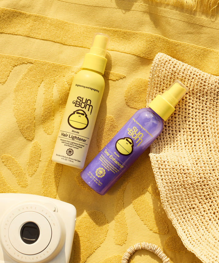 Blonde Lighten & Tone Kit featuring two spray bottles in yellow and purple, displayed on a sunny yellow towel with a straw hat and instant camera. Natural hair lightening and toning products by Sun Bum.
