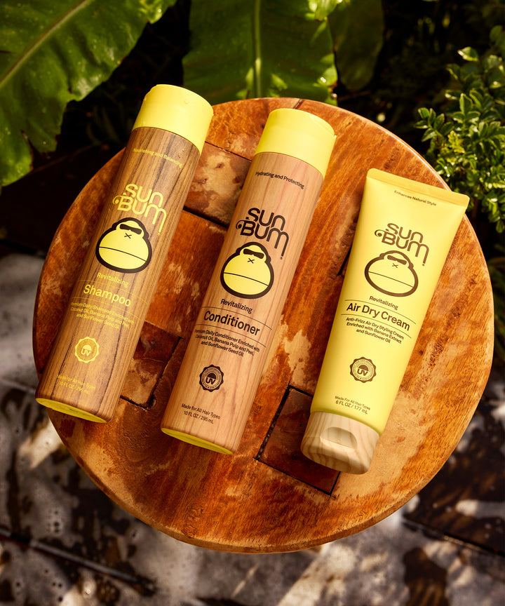 Three Sun Bum hair care products, including their Revitalizing Conditioner, displayed on a rustic wooden surface against a leafy background. The bottles feature a natural bamboo-like design with yellow caps and the iconic Sun Bum monkey logo - by Sun Bum