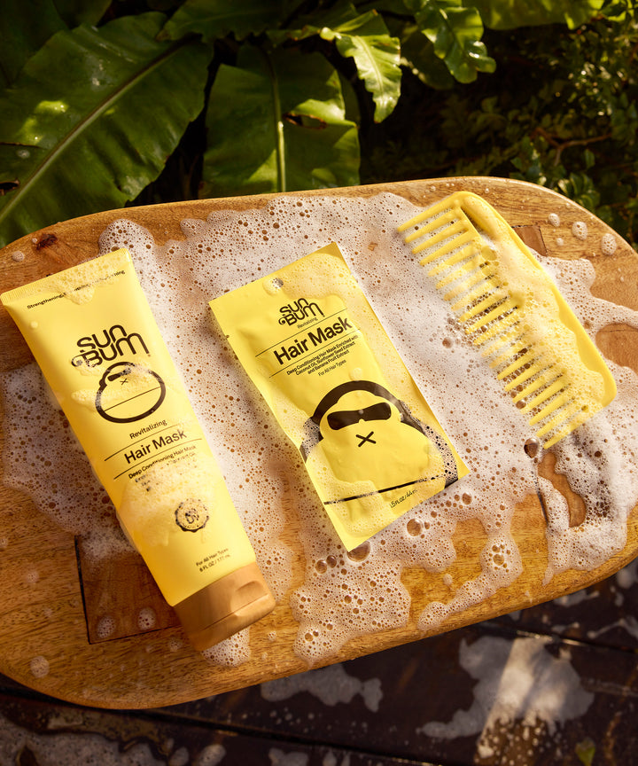 Three yellow tubes of Revitalizing Hair Mask displayed on a wet wooden surface with tropical leaves in background, featuring nourishing hair treatment products for damaged hair by Sun Bum