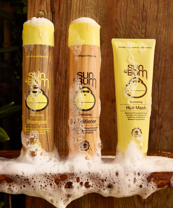 Three yellow Sun Bum hair care products, including shampoo, conditioner, and Revitalizing Hair Mask, displayed on wooden surface with foamy bubbles, showcasing complete hair care system by Sun Bum