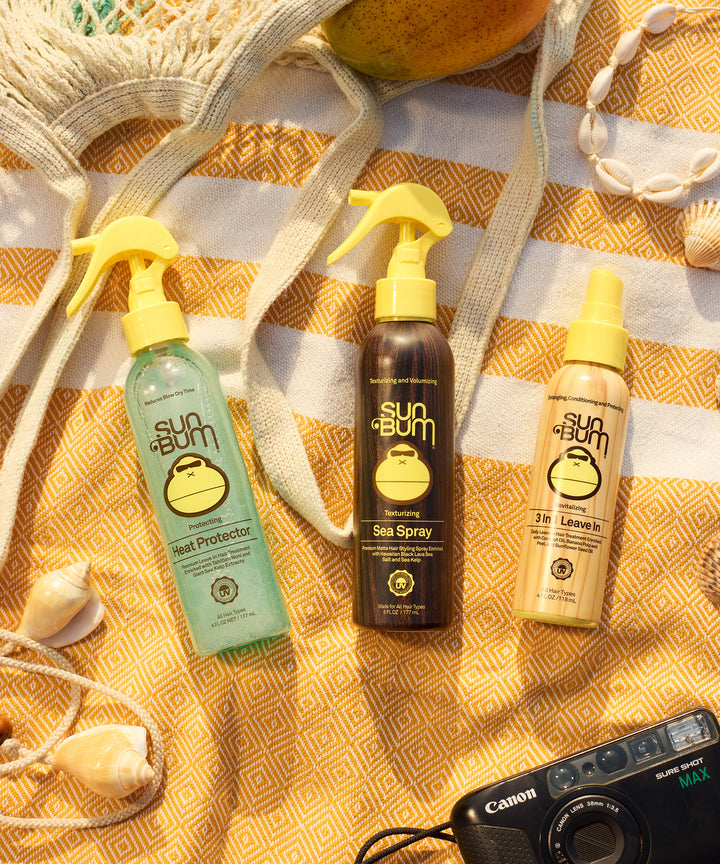 Three Sun Bum hair styling products, including Sea Spray, displayed on a yellow geometric patterned beach towel with seashells and a vintage camera, creating a summer beach-themed flatlay by Sun Bum