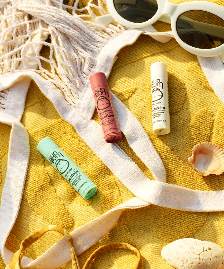 Three CocoBalm Lip Balm tubes in different colors displayed on a yellow beach towel with white sunglasses, seashells, and rope accents, creating a sunny summer vibe. CocoBalm Lip Balm 3-Pack by Sun Bum.