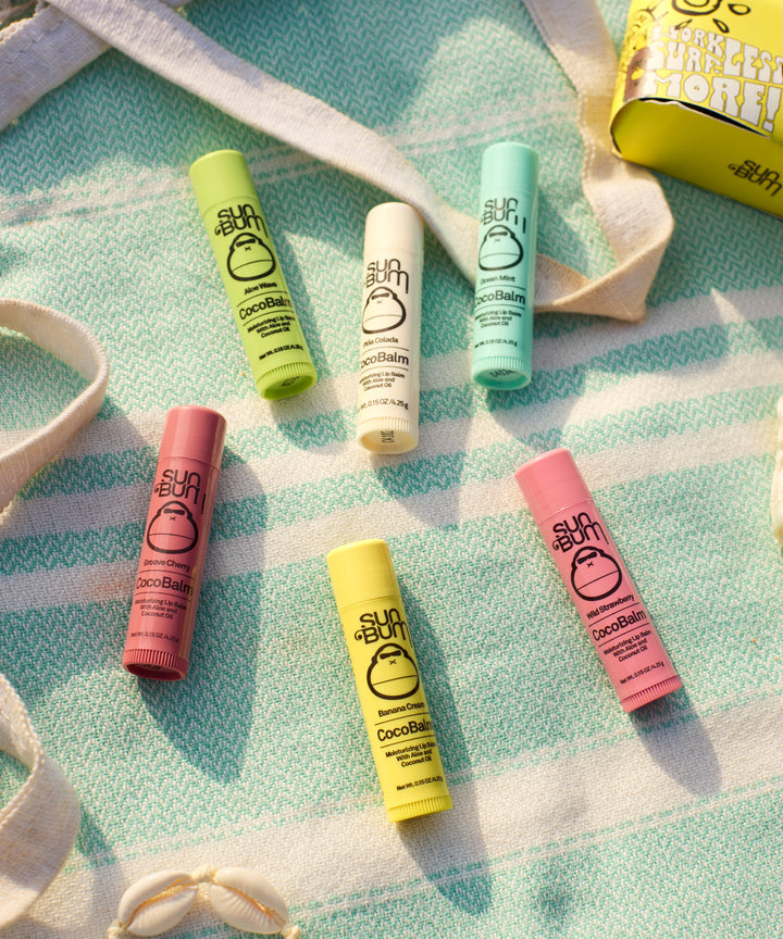Colorful CocoBalm Ocean Mint lip balms arranged on a mint green and white patterned fabric, featuring pastel tubes in pink, yellow, green, and white. Each tube displays the Sun Bum logo and CocoBalm branding against a soft, beach-inspired backdrop. By Sun Bum.