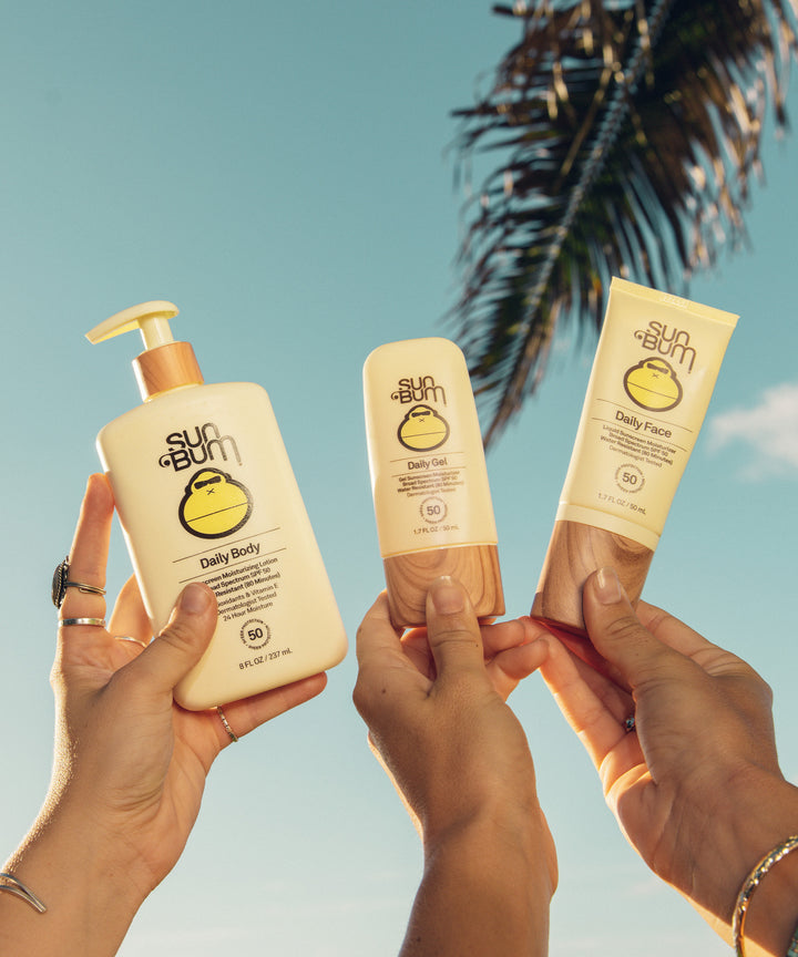 Three hands holding Sun Bum skincare products, including Daily Body Lotion, Daily 50 Face Gel, and Daily Face Sunscreen, against a sunny backdrop with a palm tree. Sun Bum.