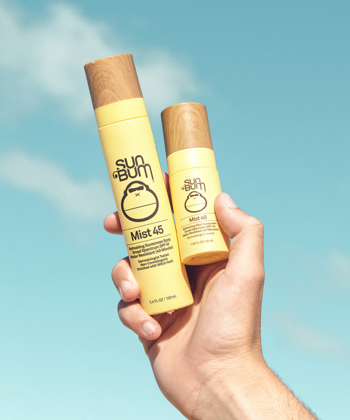 Original SPF 45 Sunscreen Face Mist by Sun Bum shown in two yellow spray bottles with wooden caps against a bright blue sky background, held up to showcase the lightweight, protective facial sunscreen mist - Sun Bum