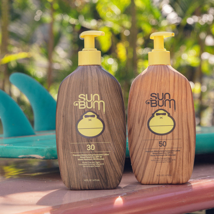 Original SPF 50 Sunscreen Lotion by Sun Bum on a sunny day, featuring a wooden-themed bottle next to a SPF 30 version, close to surfboards, Sun Bum