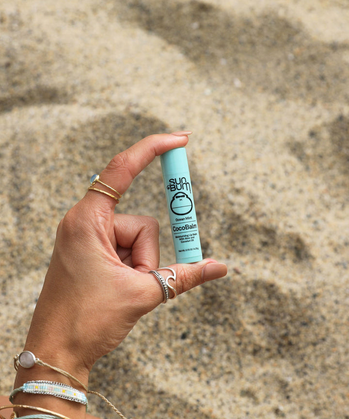 Hand holding CocoBalm Lip Balm - Ocean Mint by Sun Bum, against a sandy beach background.