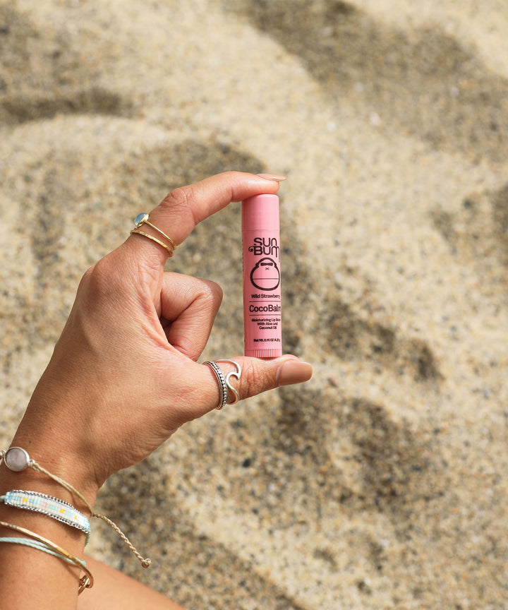 Hand holding a pink tube of CocoBalm Lip Balm - Wild Strawberry flavor, Sun Bum, against a sandy beach background.
