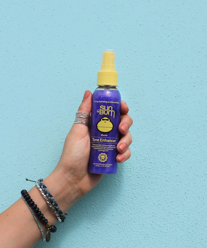 Hand holding a bottle of Sun Bum Blonde Tone Enhancer against a light blue background.