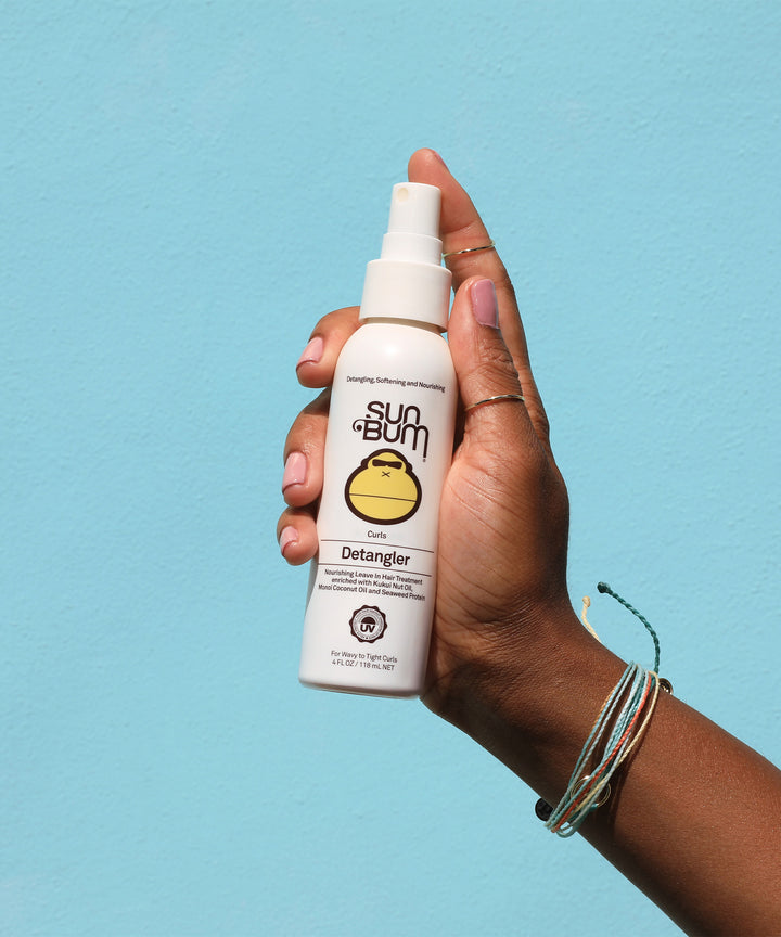 Hand holding a bottle of Curls Detangler spray, made with Kukui Nut Oil and Monoi Coconut Oil to soften, tame, and condition curly hair, by Sun Bum.