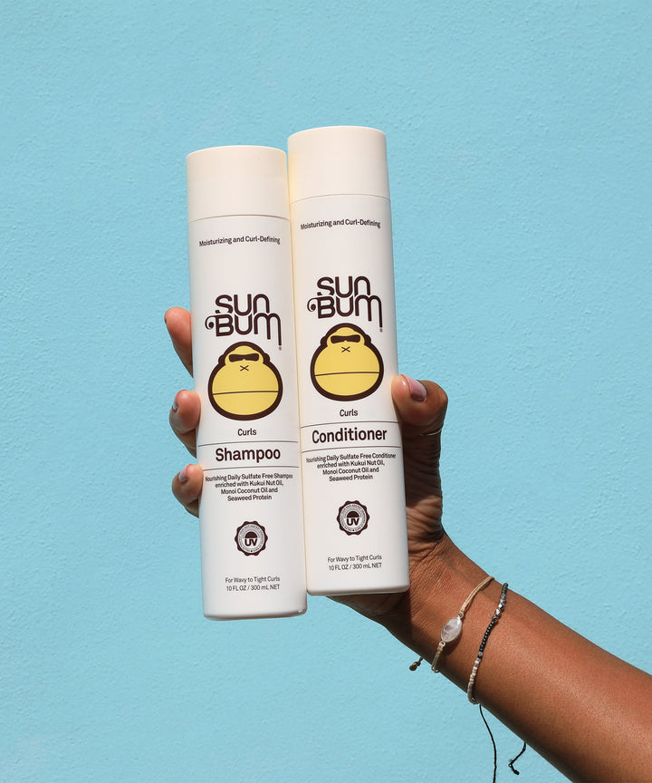 A hand holding two bottles of Sun Bum Curls Shampoo and Conditioner, featuring Kukui Nut Oil and Monoi Coconut Oil for curly hair care. Sun Bum.