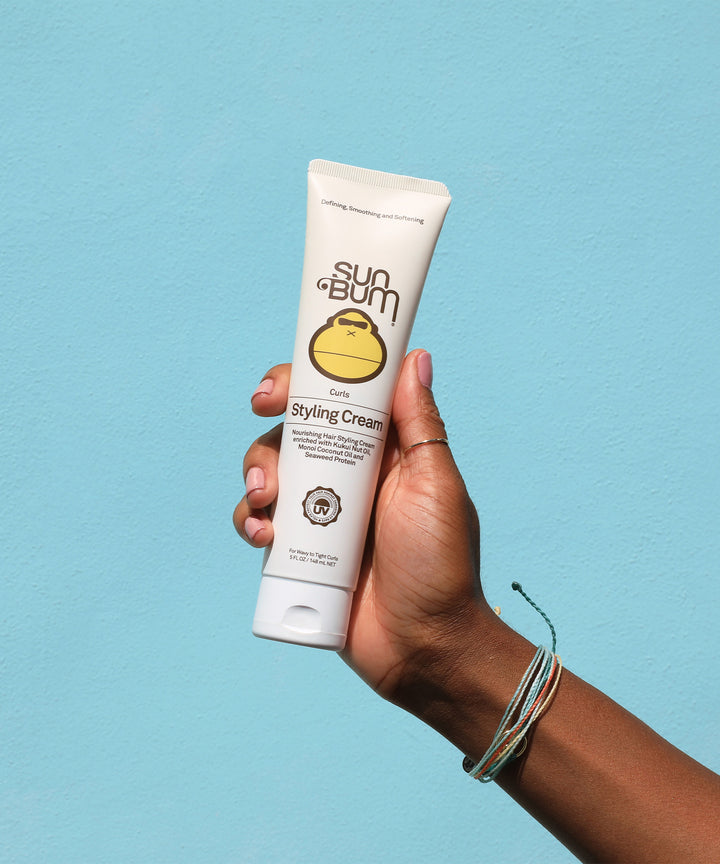 Person holding a tube of Sun Bum Curls Styling Cream with Kukui Nut Oil and Monoi Coconut Oil against a light blue background, Sun Bum
