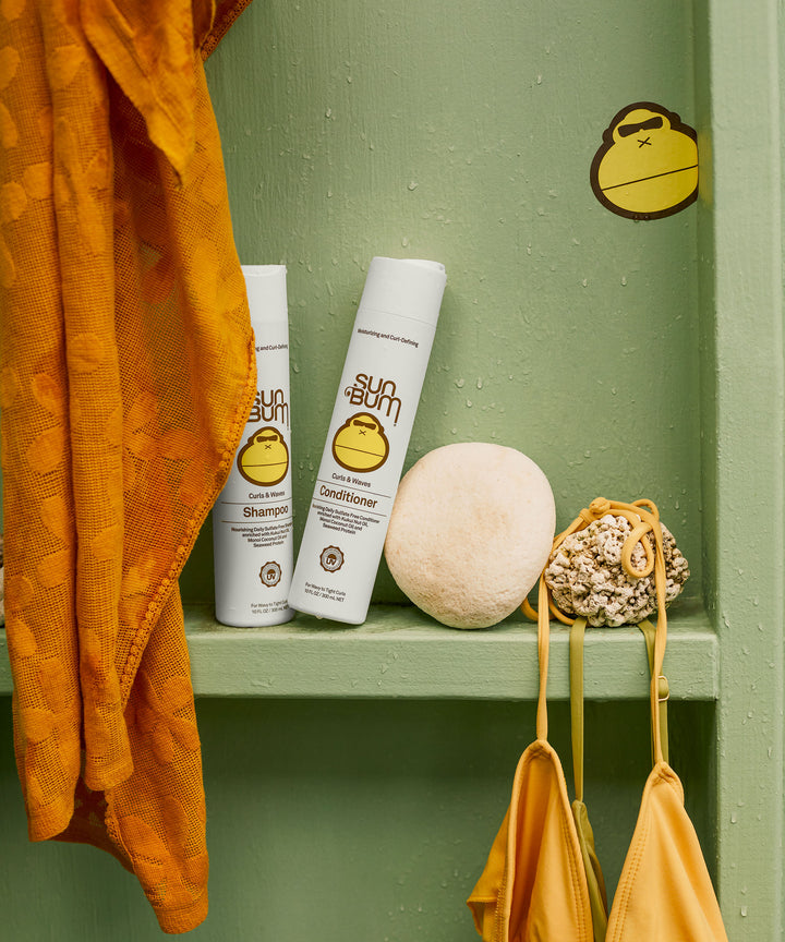 Sun Bum Curls Conditioner and Shampoo on a green shelf, paired with a yellow towel, sponge, and yellow bikini top, Sun Bum
