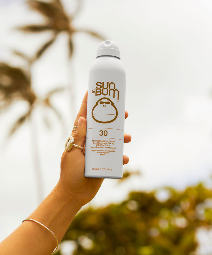 Hand holding Mineral SPF 30 Sunscreen Spray by Sun Bum, with a backdrop of palm trees and a cloudy sky, showcasing the zinc-based, fragrance-free, B.O.V technology sunscreen.