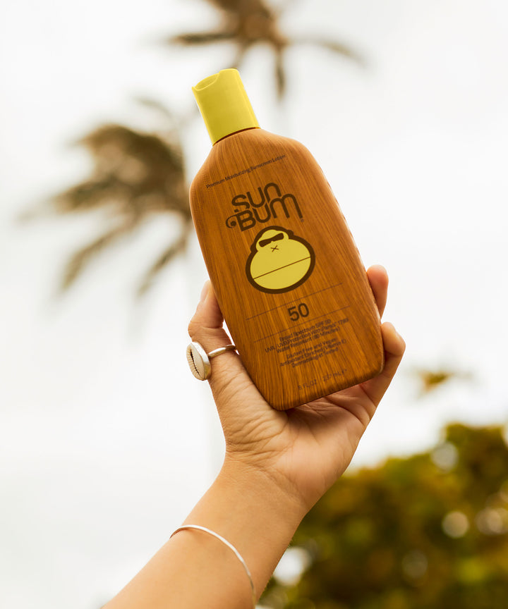Hand holding a bottle of Original SPF 50 Sunscreen Lotion with palm trees in the background, Sun Bum.