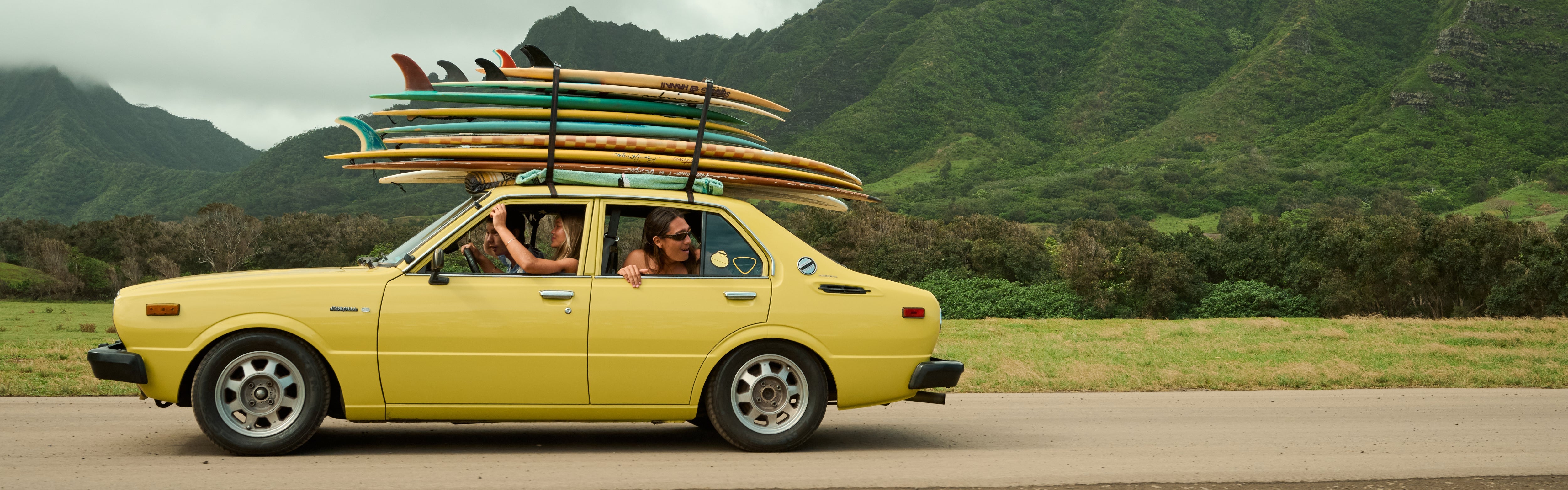 Friends driving a yellow car stacked with surfboards through lush mountains, featuring the SB After Sun Care Collection – Sun Bum.