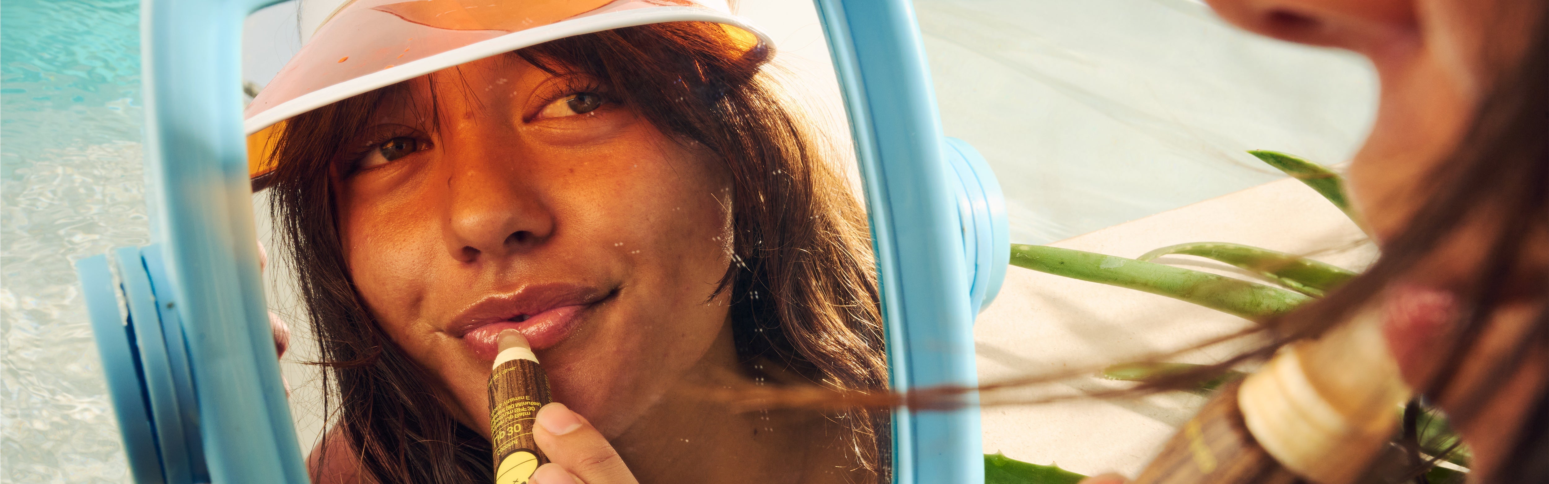 Woman applying SPF lip balm in a mirror reflection while sunbathing, featuring the SB Lip Care Collection – Sun Bum.