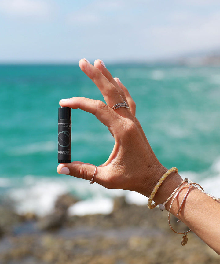 Hand holding Signature SPF 30 Sunscreen Lip Balm with ocean background, perfect for sun protection outdoors. Sun Bum.