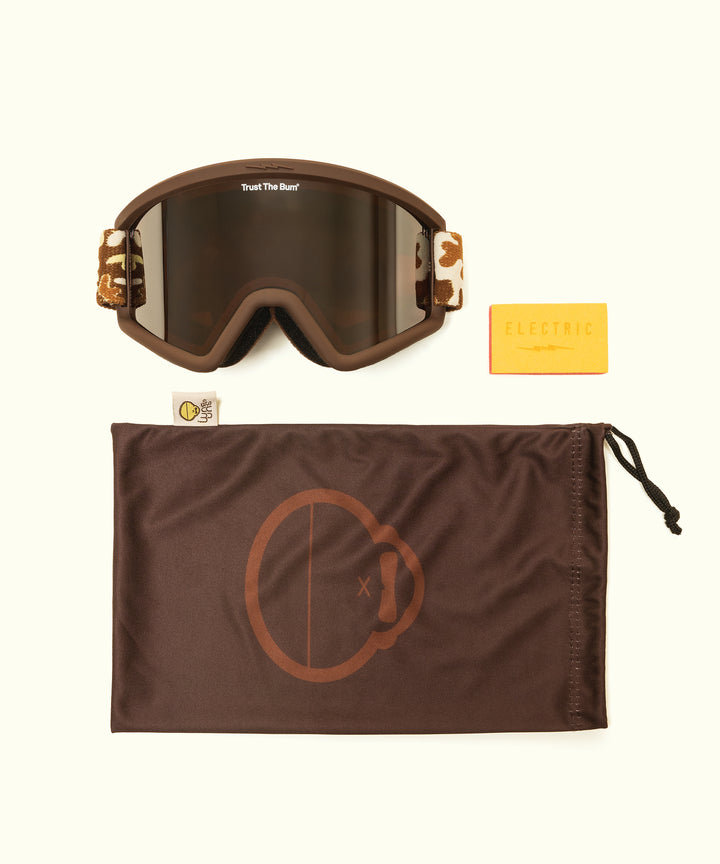 Brown snow goggles with tinted lenses shown with branded drawstring bag and yellow accessory card. Sun Bum X Electric Goggles featuring protective eye wear design for mountain activities. - Sun Bum