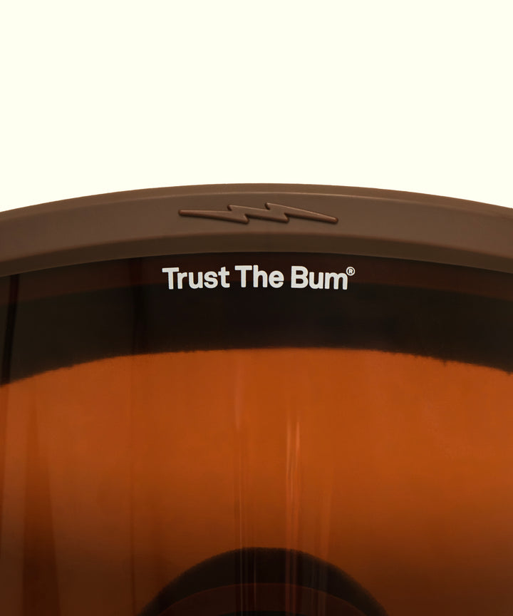 Sun Bum X Electric Goggles featuring a brown-tinted lens with orange gradient and Trust The Bum branding on the frame. Premium snow goggles designed for mountain protection and style. By Sun Bum.